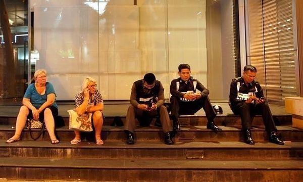 Khách du lịch ngồi cạnh cảnh sát Thái Lan trước cửa một trung tâm mua sắm gần hiện trường vụ nổ. Ảnh: Guardian